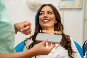 Woman sitting in dentist’s chair smiling while staff member holds veneers in foreground.